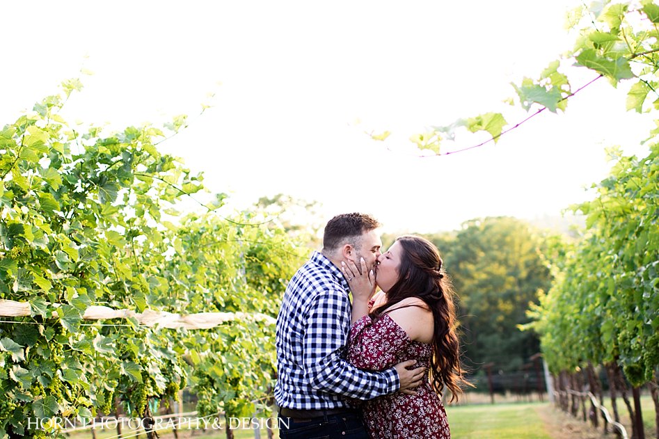 engaged couple kissing and embracing blue and white checked shirt red floral dress in vineyard Metro Atlanta  husband and wife team horn photography and design